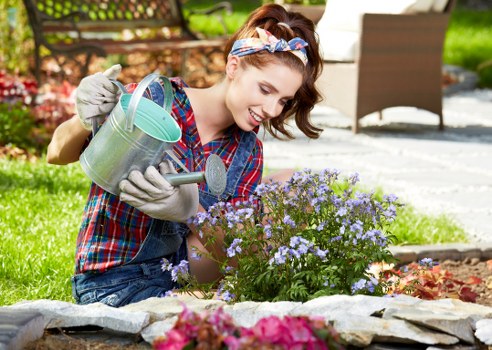 A young woman with a cheerful expression is kneeling in a well-maintained garden, watering a flower bed filled with small, purple flowering plants using a green metal watering can. She is wearing a colorful plaid shirt, grey gardening gloves, and a striped headband. The garden features a neatly edged flower bed bordered with flat stones, with vibrant pink flowers in the foreground and lush green grass around. In the background, there is a paved patio area with outdoor furniture, including a table and chairs, suggesting a private outdoor space in Queens Park. The scene is captured on a bright, sunny day with natural light illuminating the garden and the woman's activity, reflecting typical garden maintenance practices that a professional gardening service, such as Gardening Services Queens Park, might undertake in the area. The setting emphasizes a vibrant, tidy, and inviting outdoor environment suitable for outdoor leisure and gardening care.