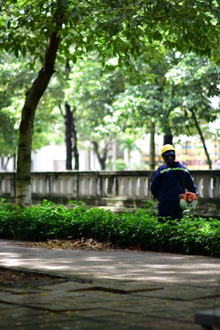 A gardener wearing a navy blue uniform with reflective yellow stripes and a protective yellow helmet is using a portable hedge trimmer to trim a neatly maintained hedge along a paved pathway in a park