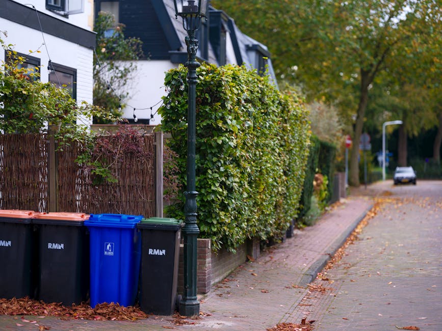 A black wheeled garden waste bin positioned on a grassy lawn in a garden setting, filled with assorted garden debris including dried ornamental grasses, green leaves, and cut branches. The background 